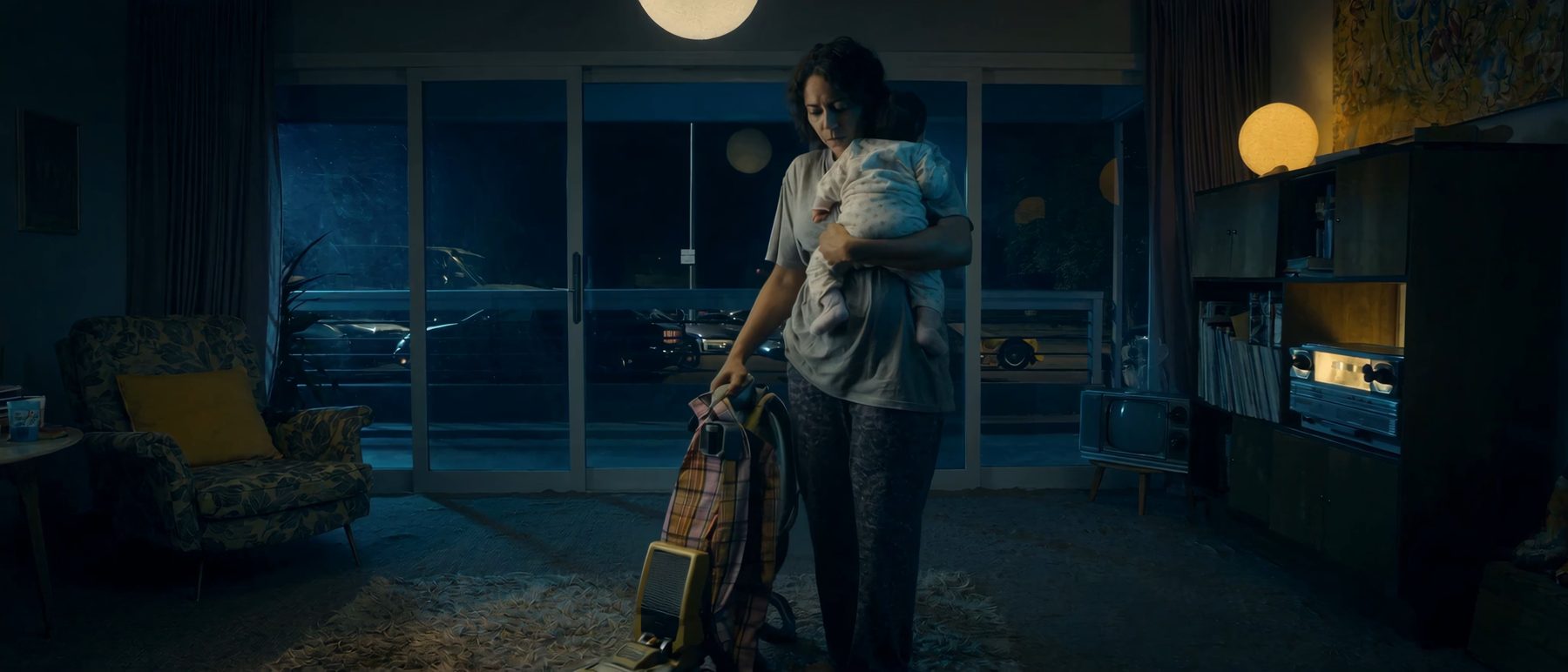 An exhausted mother vacuuming at night, baby on her hip