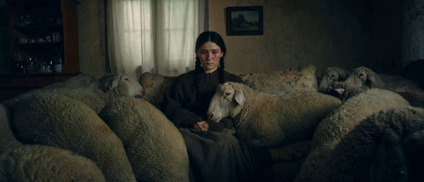 A woman on a worn floral sofa, sheep pressed close around her