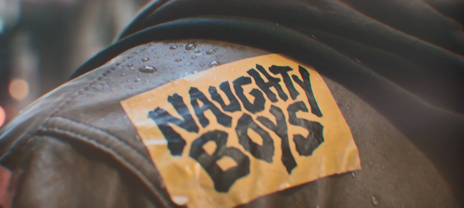 Macro shot of a yellow Naughty Boys patch, its hand-drawn lettering stitched onto a rain-soaked leather jacket