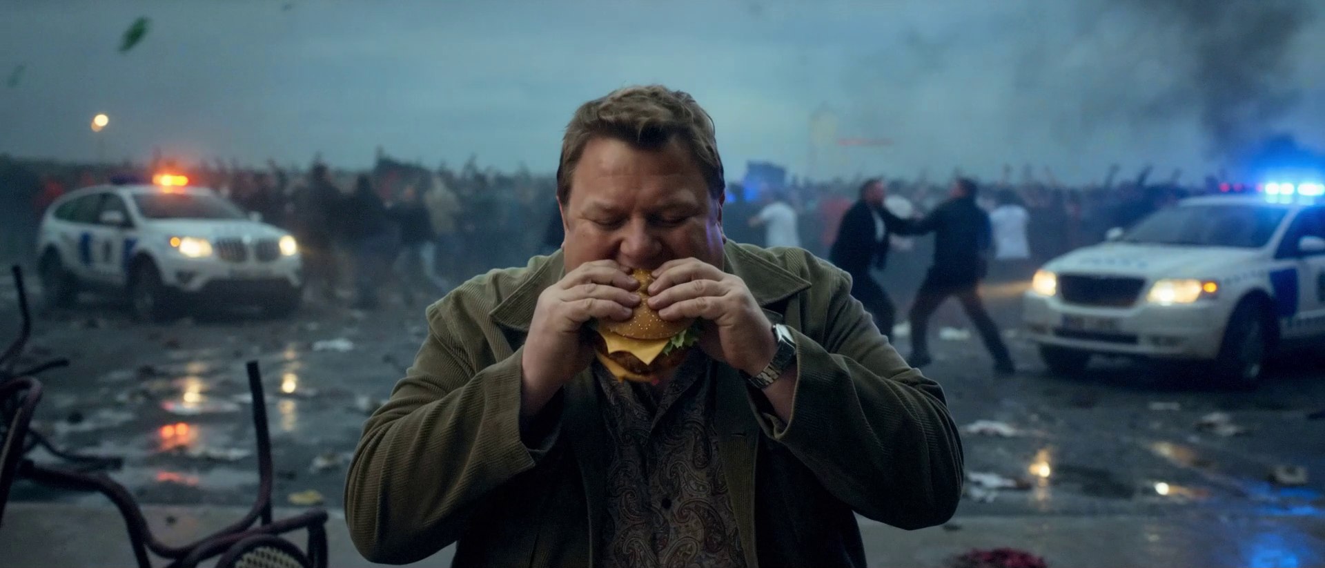 A man calmly eats a cheeseburger in the middle of a street riot, police cars and a charging crowd behind him