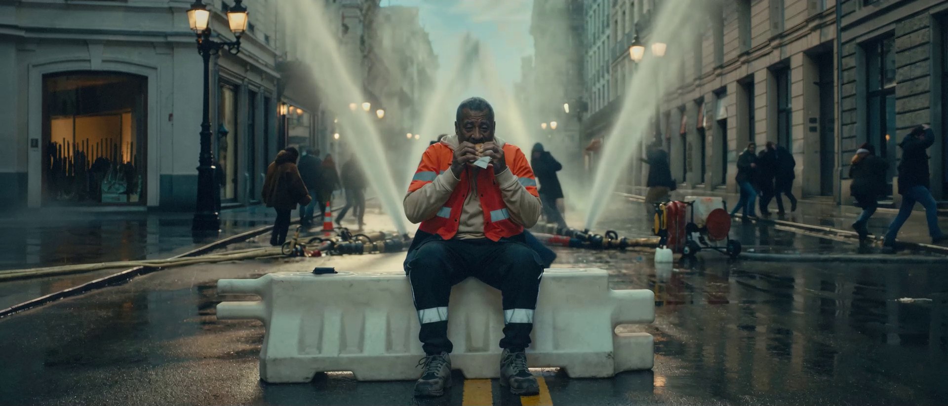 A man in a hi-vis vest eats on a road barrier as water cannons blast down a European street around him