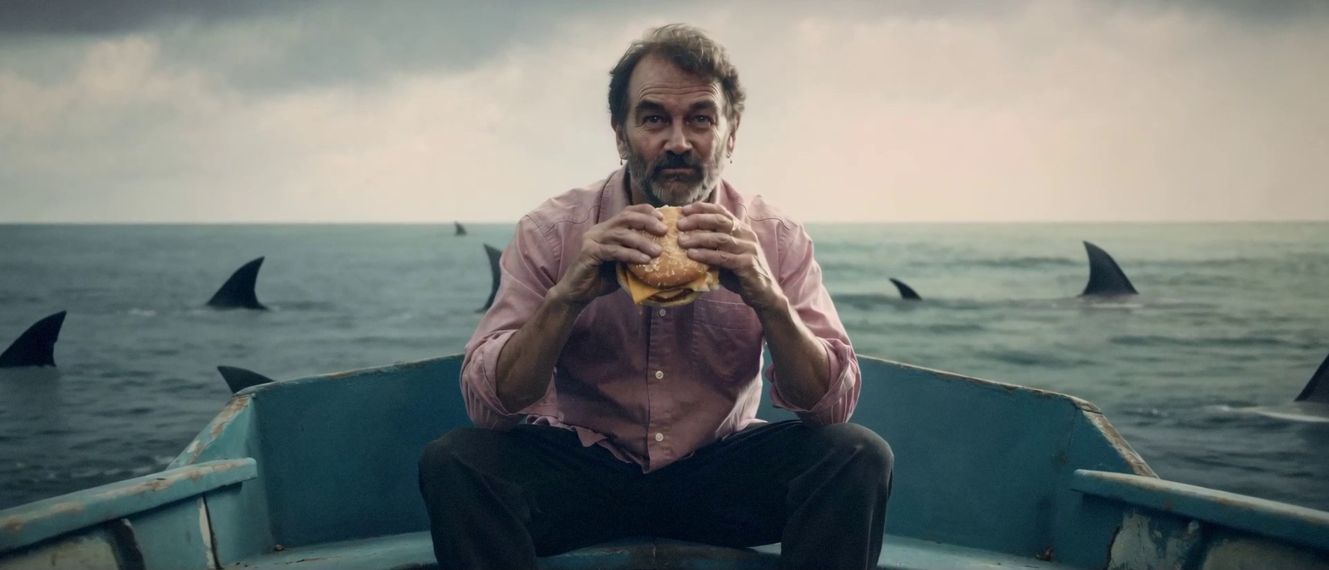 A man eats a burger while sitting calmly in a small boat ringed by circling shark fins