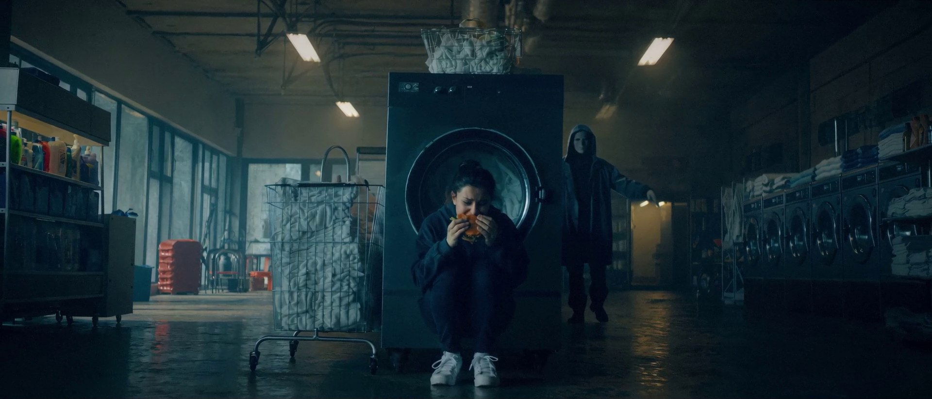 A woman crouches eating a burger against a laundromat dryer as a hooded figure creeps up behind her