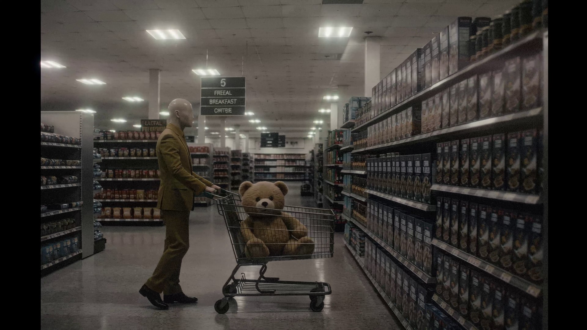 The man pushes a supermarket trolley down an empty aisle with a giant teddy bear riding in it
