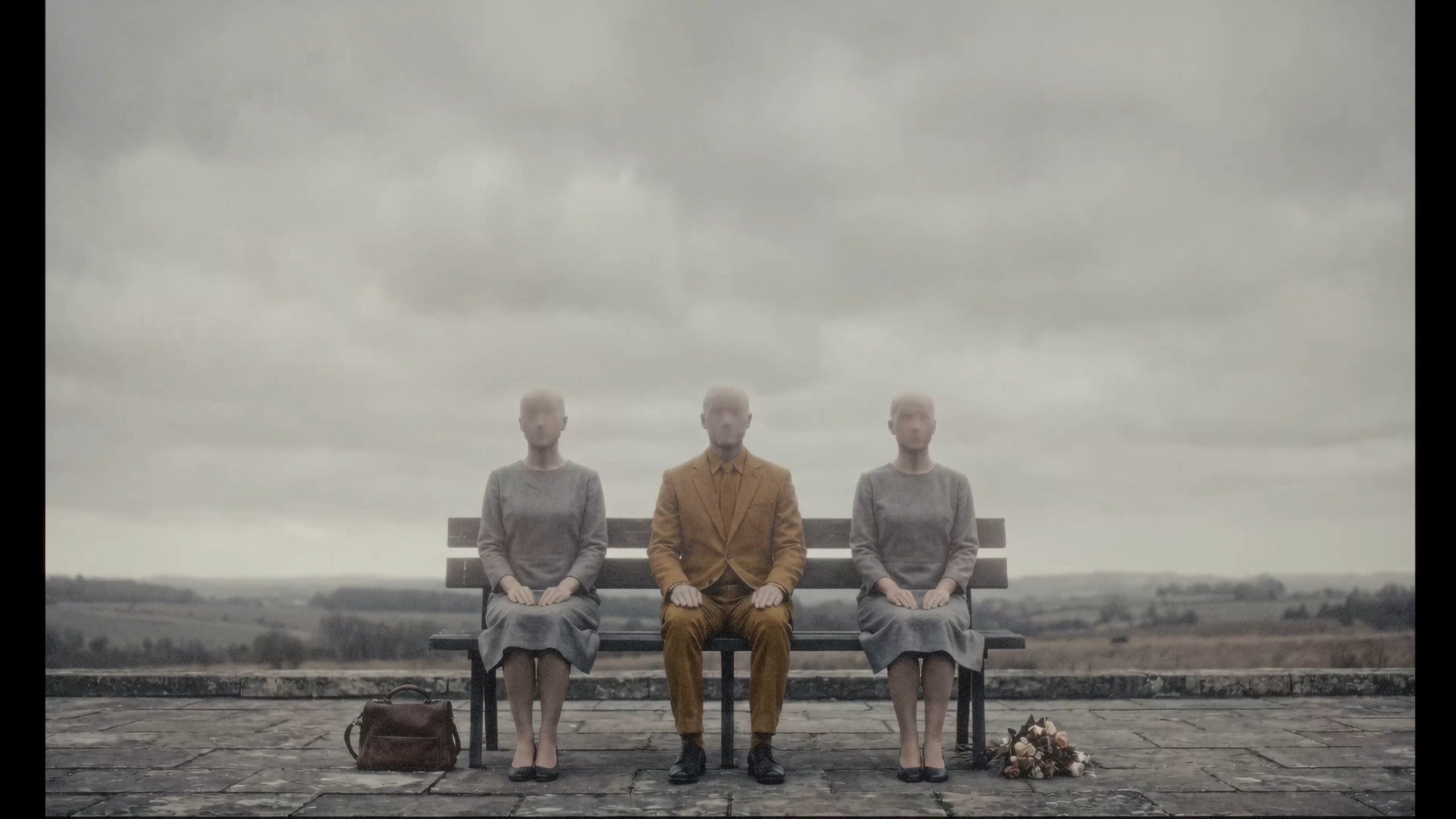 Three faceless figures in muted grey clothing and one in a mustard suit sit on a bench overlooking a wide, overcast countryside landscape