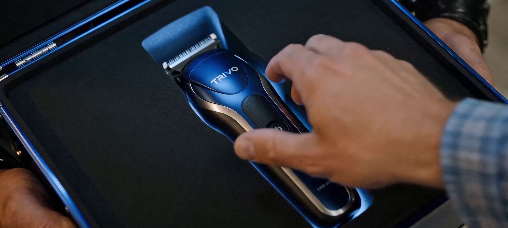 Close-up of the blue and gunmetal Trivo trimmer seated in its foam-lined case, a hand reaching for it