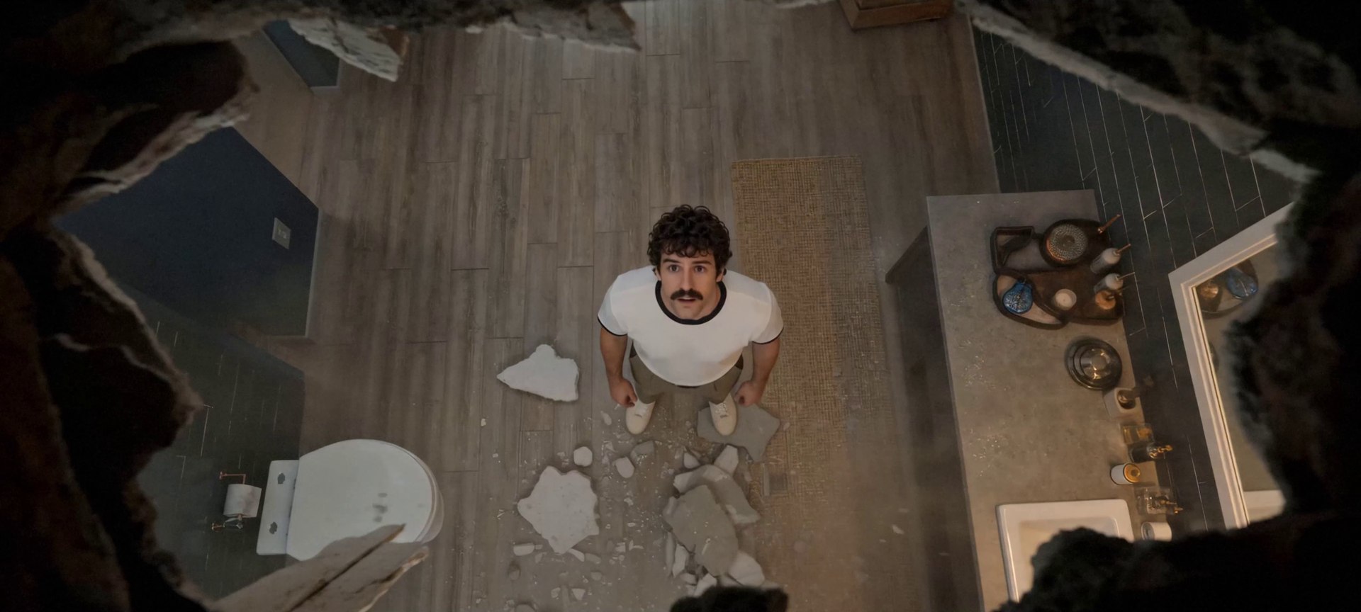 Overhead shot of a man standing in his bathroom looking up through a jagged hole smashed in the ceiling