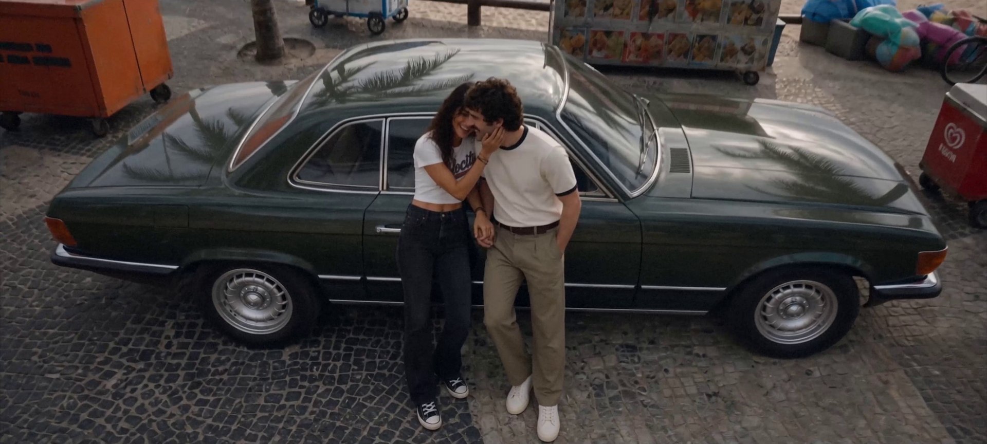 A couple lean against a vintage green coupe on a cobbled seaside street, foreheads together, laughing